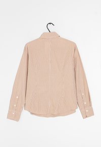 Camisa de cuadros en tonos marrones y blancos con collar, mangas largas, dobladillo redondeado y acentos de botón blanco en los puños, colgando de un gancho negro.