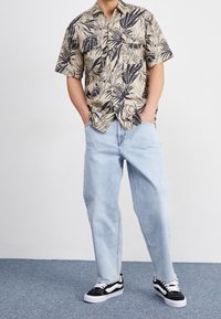 Beżowa koszula z krótkim rękawem w czarny kwiatowy wzór, zestawiona z jasnoniebieskimi luźnymi dżinsami i czarno-białymi trampkami.