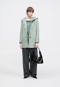 Lysegrønn kåpe i ullblanding med hette og knappedetaljer, brukt over en blågrønn topp og grå vidbukser; holder en svart veske i lakkeret skinn.