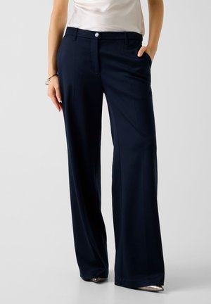 Marineblaue Hosen mit weitem Bein und glatter Textur, ausgestattet mit einer vorderen Knopfleiste und seitlichen Taschen, kombiniert mit einem enganliegenden Oberteil.