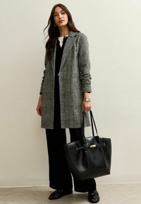 Blazer gris de cuadros largo con solapas de pico, combinado con una blusa y pantalones negros. Sosteniendo un bolso tote de cuero negro con un acento dorado.