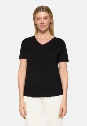 Frau trägt ein schwarzes T-Shirt mit V-Ausschnitt, das ein gepunktetes Blattmuster und den Schriftzug "SUNSET" zeigt, kombiniert mit weißen Hosen, stehend vor einem schlichten Hintergrund.