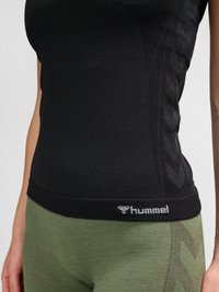 Close-up van een persoon die een zwarte mouwloze top en groene leggings draagt, beide met subtiele chevronpatronen en een zichtbaar "hummel"-logo.