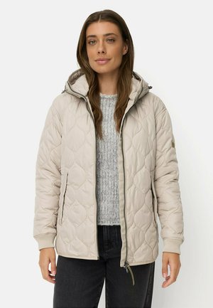 Kvinde iført beige quiltet jakke med hætte over grå strikket sweater og sorte bukser, står foran en ensfarvet baggrund.