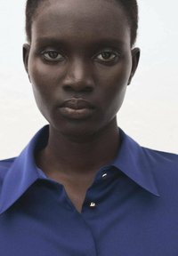 Blue collared shirt with gold button accents, smooth texture, close-up of face, short hair, neutral background.