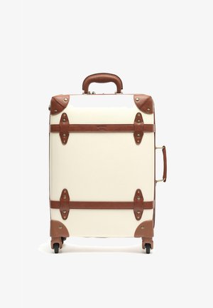 Valigia rigida color crema con dettagli in pelle marrone. Dispone di una maniglia superiore, cinghie frontali, hardware in metallo e quattro ruote.
