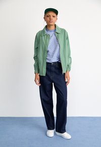 Veste bomber verte sur une chemise rayée bleue et blanche, associée à un pantalon ample bleu foncé et des baskets blanches, portée avec une casquette verte.