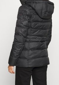 Chaqueta de plumas negra con capucha, que presenta un diseño acolchado con costuras, cierre de cremallera y un logo circular en el hombro izquierdo.