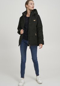 Giacca nera con zip e cappuccio, caratterizzata da dettagli color sabbia e tasche, indossata sopra una maglietta blu navy e abbinata a jeans blu e sneaker bianche.