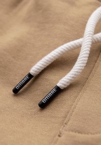 Tessuto beige con cordoncini bianchi intrecciati, punte nere etichettate "OMBRE" e cuciture visibili vicino al bordo.