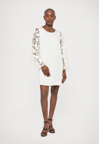 Vestido blanco con mangas largas y transparentes de campana adornadas con aplicaciones florales. La tela es suave, con un corte relajado y escote redondo.
