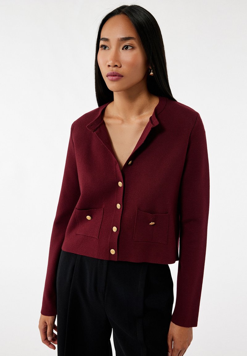 Burgunderfarbene, kurz geschnittene Strickjacke aus weichem Material mit Kragen, vorderem Knopfverschluss, zwei vorderen Taschen und Knöpfen in Gold-Optik.