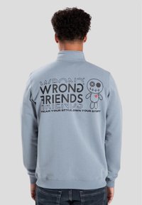 Ljust blå sweatshirt med hög krage, prydd med svart text och en karaktärsgrafik på baksidan. Mjuk textur, avslappnad passform.