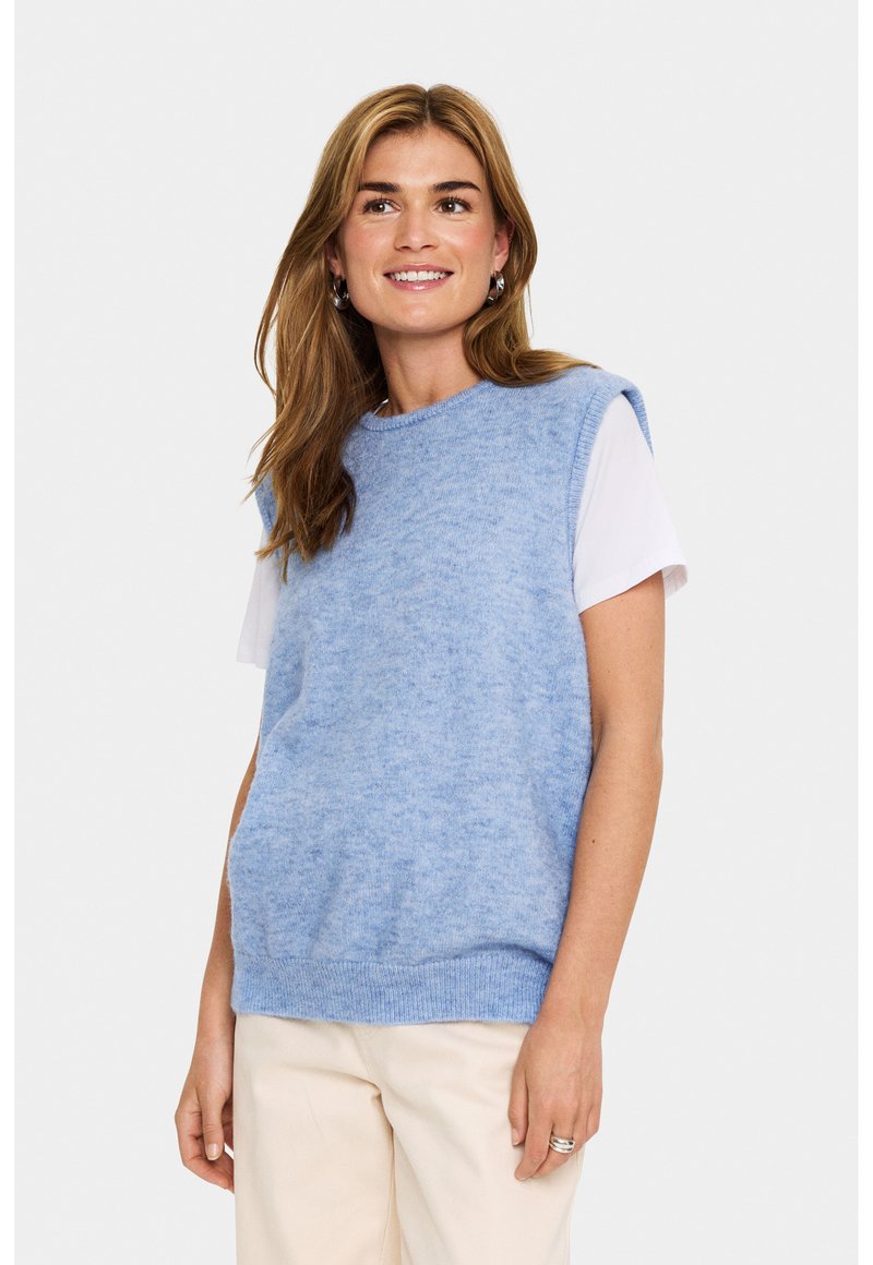 Pulover tricotat fără mâneci, de culoare albastră, cu o textură moale, decolteu rotund și tiv cu dungi, purtat peste un tricou alb. Pantaloni bej deschis.