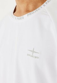 T-shirt bianco a maniche corte con scollatura rotonda. Presenta un logo grigio ricamato sul petto e un colletto a fantasia con la scritta "PROJECT X PARIS."