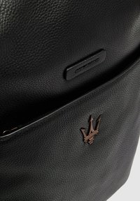 Mochila de cuero negro con acabado texturizado, parche de logo sutil y un adorno de emblema en bronce. Incluye un bolsillo frontal con cremallera.