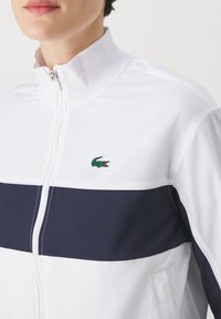 Chaqueta blanca con cremallera y rayas horizontales en azul marino, que presenta un pequeño logo de cocodrilo verde; material suave y ligero con un cuello alto.