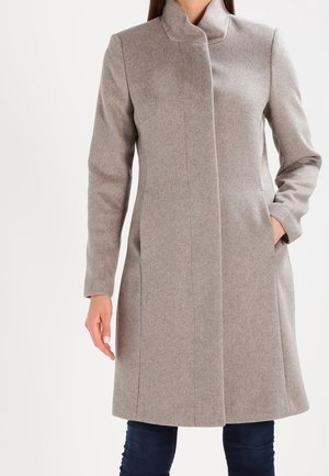 Kobieta w sięgającym kolan, jasnoszarym wełnianym płaszczu z kołnierzem typu stójka, bocznymi kieszeniami i długimi rękawami, nosząca ciemne spodnie.