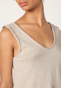 Naine, kes kannab varrukateta beeži toppi, millel on töötlemata servadega käeaugud ja kaelus, mis paljastab tema kaela, õla ja ülakeha.