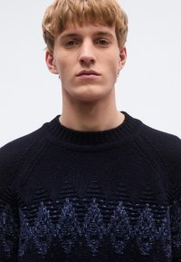 Sötét tengerészkék kötött pulóver, alsó félrészen texturált, geometrikus mintával kék árnyalatokban. Bordázott környakú és raglán ujjú.