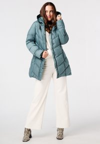 Teal pufferjack met een capuchon, voorzien van een diamantpatroon en een sluiting aan de voorkant. Draagt over een witte trui en wijde broek.