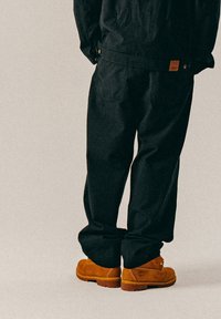 Mustat teksturoitu housut, joissa on löysä Fit ja näkyvät taskut, yhdistettynä ruskeisiin mokkanahkakenkiin, joissa on pieni merkintä sivussa.