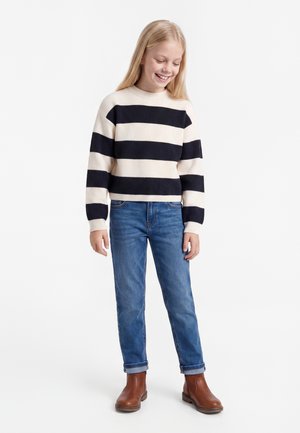 Junges Mädchen lächelt, trägt einen schwarz-weiß gestreiften Pullover, blaue Jeans und braune Stiefeletten, steht vor weißem Hintergrund.