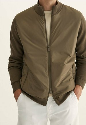 Mand iført olivengrøn lynlåstrøje med ribstrikkede ærmer og krave, hænderne i lommerne, kombineret med hvide bukser.