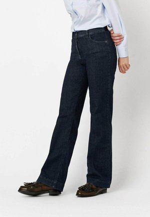 Person trägt dunkelblaue, hoch taillierte Jeans mit geradem Bein, hellblaues Hemd mit Knöpfen und braune Loafers mit Quasten und steht mit einer Hand hinter dem Rücken.