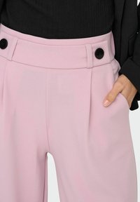 Pink tailored trousers with a high waistband, featuring black buttons, side pockets, and a smooth, lightweight fabric texture.
