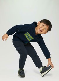 Navyblauer Kapuzenpullover mit dem Aufdruck "STAY COOL", kombiniert mit dunklen Cargo-Hosen. Das Kind trägt schwarze Sneaker mit grünen Akzenten und nimmt eine verspielte Pose ein.