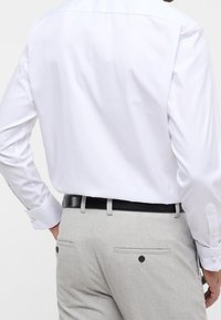 Valkoinen, pitkähihainen paita, jossa on sileä pinta, yhdistettynä vaaleanharmaisiin kuvioituihin housuihin, joissa on kaksi takataskua ja musta vyö.