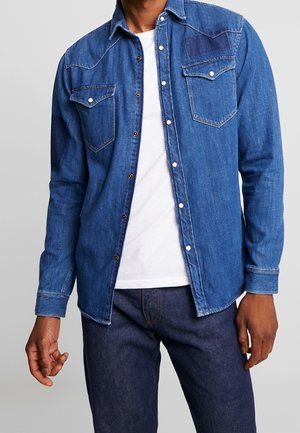 Mężczyzna w niebieskiej dżinsowej koszuli zapinanej na guziki, na białej koszulce i ciemnoniebieskich dżinsach, stojący na jasnym, jednolitym tle.