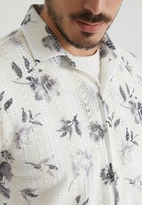 Chemise à manches courtes en tissu en filet blanc avec un imprimé floral noir, une texture subtile et un col classique.