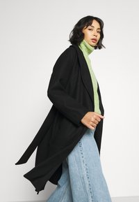 Black long coat with open front design, worn over a light green turtleneck. Features loose, flowing sleeves and a relaxed fit. Light blue jeans.