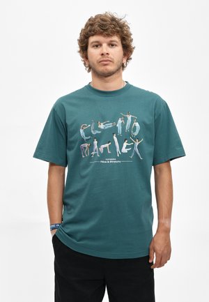 Mann mit lockigem Haar, der ein türkisfarbenes T-Shirt trägt und mehrere kleine Tänzer in verschiedenen Posen zeigt, die das Wort "CLEOPHANTRIK." formen