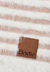 Neulottu kangas, jossa vuorottelevat valkoiset ja vaaleanpunaiset raidat, ja pehmeä tekstuuri. Ruskea nahkalappu, jossa on kirjailtuna "Elodie", on näkyvissä.