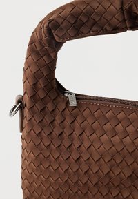 Sac à main marron tissé avec une poignée rembourrée, une fermeture éclair et un accent métallique. Présente un motif texturé et une forme incurvée.