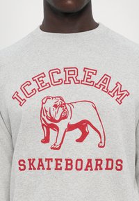 Grå sweatshirt med et rødt bulldoggrafik, hvor "ICECREAM" står ovenfor og "SKATEBOARDS" står nedenfor i fed rødt skrift. Standard pasform.