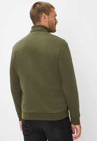 Felpa verde con collo alto e texture liscia, con logo sul collo e polsini e orlo a coste. Indossata con pantaloni neri.
