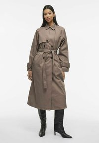 Lungo trench coat marrone con vita cintrata, bottoni doppi e ampi rever. Presenta maniche lunghe con polsini regolabili.