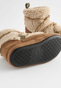 Tarn- und beige gefütterte Stiefel mit Gummisohlen, ausgestattet mit gekreuzten Riemen und einer Zuglasche, die ein strukturiertes Sohlenmuster betont.