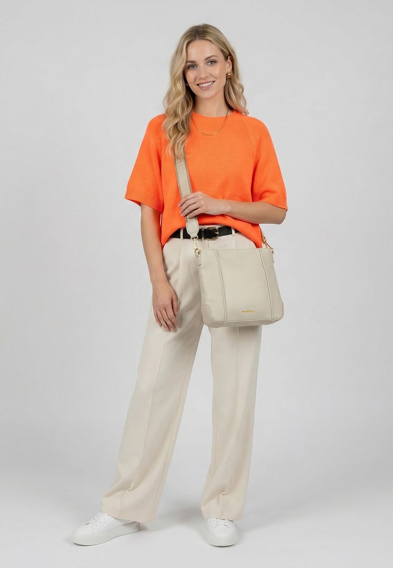 Femme portant un pull à manches courtes orange vif, un pantalon large beige, des baskets blanches, tenant un sac bandoulière beige, souriante devant un fond uni.