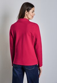 Fuksia pitkähihaine paita, jossa on kaulus ja yksivärinen pinta. Takapuoli on sileä, rento malli ja erittäin vähäiset ommellut yksityiskohdat. Yhdistetty tummiin farkkuihin.