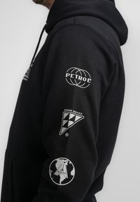 Felpa nera con cappuccio caratterizzata da più loghi: "PETROL" in bianco, un emblema triangolare "DENIM DRIVEN" e un'icona del globo "ALASKA" sulle maniche.