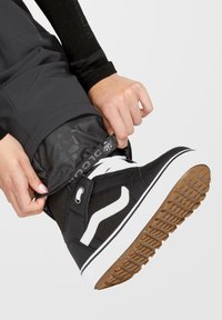 Schwarz-weiße Sneaker mit einer strukturierten Gummisohle und einem flexiblen Obermaterial. Mit Branding und Schnürdesign, positioniert in der Nähe eines Hosenbunds.
