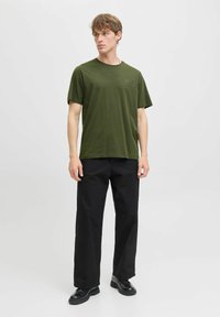 Camiseta verde oliva con cuello redondo y mangas cortas, combinada con pantalones anchos negros y zapatos negros. Diseño sencillo, sin patrones.