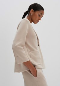 Blazer su misura beige chiaro con maniche a tre quarti, chiusura con un singolo bottone e tasche frontali, realizzato in un tessuto liscio e morbido.