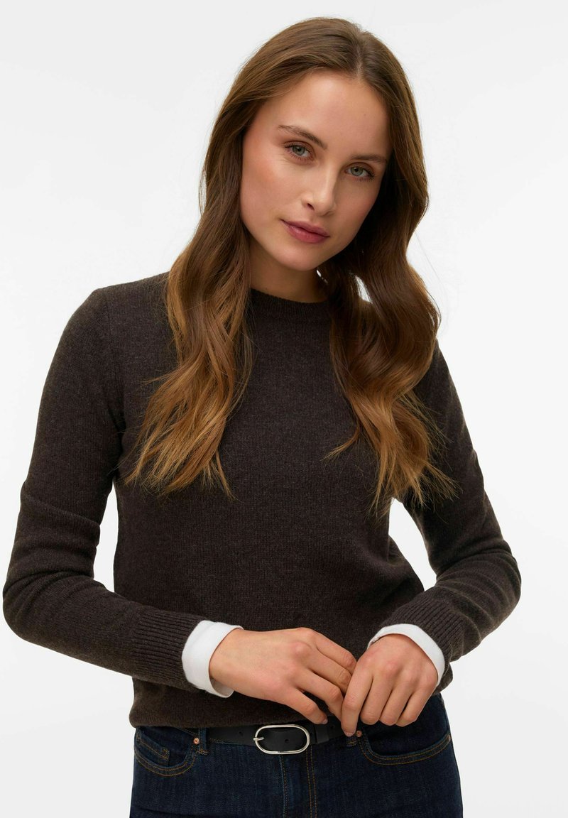 Brauner Strickpullover mit rundem Ausschnitt und gerippten Bündchen, getragen über einem weißen Hemd mit Kragen, kombiniert mit dunklen Denim-Jeans.