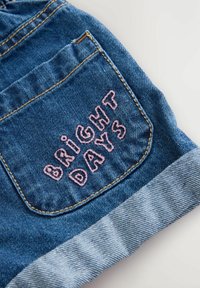 Szorty dżinsowe z podwiniętym dołem, z tylną kieszenią haftowaną frazą "BRIGHT DAYS" w kolorze różowym, na niebieskim tle.